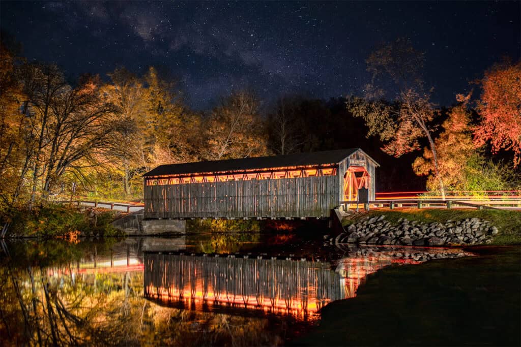 Historic covered bridge under starry sky with reflections on water.