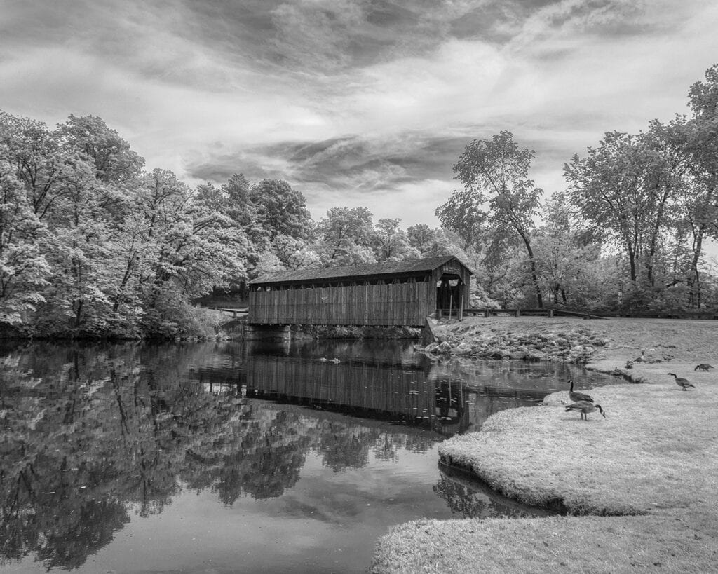 Old covered bridge over river with trees, in black and white.